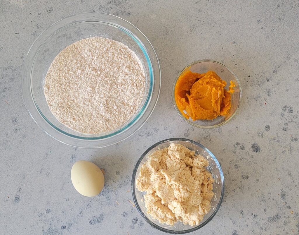 One glass bowl of flour, one glass bowl of pureed pumpkin, one glass bowl of shredded tuna and one egg on a grey background.